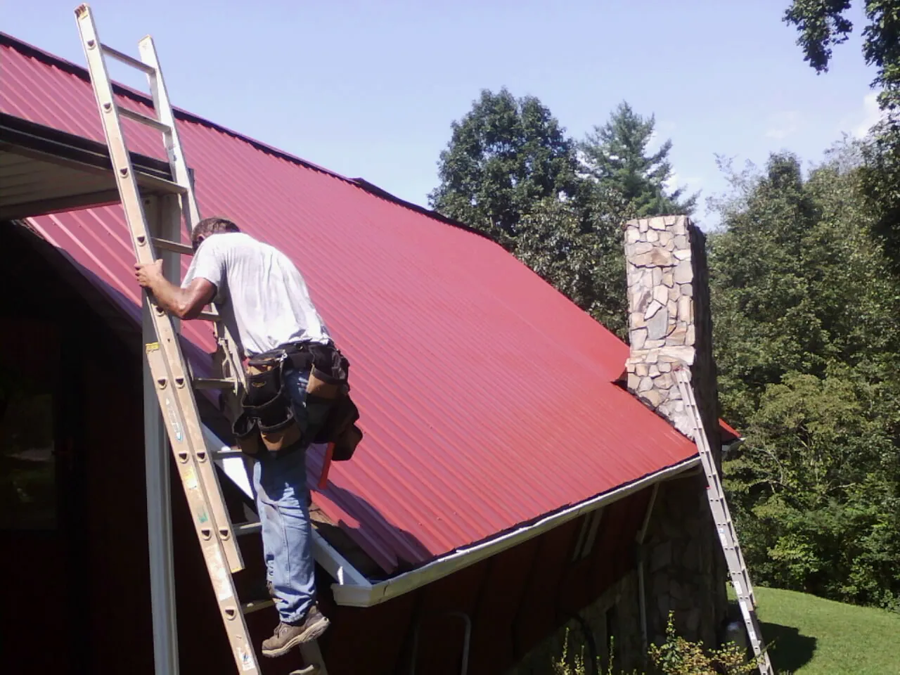Roofer on a ladder beside a red metal roof and stone chimney — Soffit & Fascia Repair by Roofing Brothers in Suquamish
